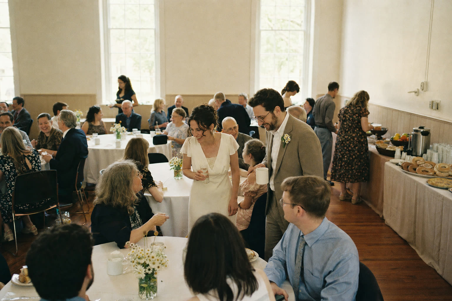 Guests at round tables in a bright community hall for a wedding brunch with a buffet along the wall